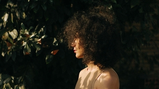 Deena Abdelwahed is framed by greenery in a garden. There are leafy plants surrounding her. Deena is wearing a sand coloured vest top and is looking to the left. She wears her curly hair loose and down, covering some of her face