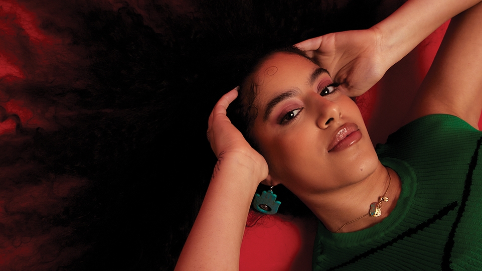 Press shot of Nooriyah lying on a red floor. Her curly hair is sprawled across the floor and she is holding either side of her head. She's wearing a green sleeveless top and green earrings