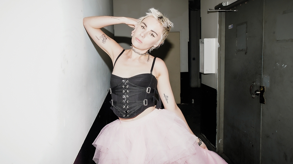 Photo of AK Sports posing in an industrial-looking hallway. They're wearing a black corset top and a flowing pink skirt. Their hair is cropped short and blonde and their hand is resting on their head with their elbow pointing out 