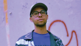 DJ Swisha standing in front of a lilac coloured wall with graffiti