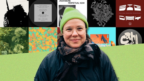 Doc Sleep smiling in a dark blue rain coat and bright green hat  in front of a selection of artwork from her Selections