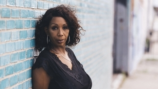 Photo of DJ Minx leaning against a blue wall and wearing a black v-neck top