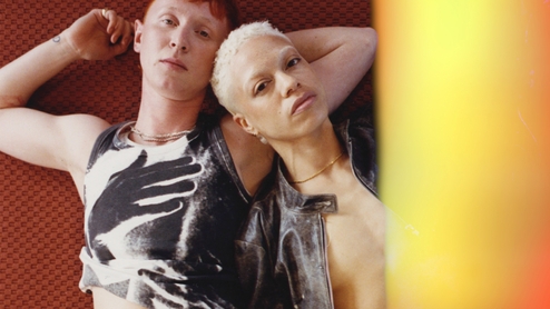 Photo of Sarah Nimmo and Reva Gauntlett lying side by side on a rusty red carpet. An orange/yellow light effect covers the right side of the imge