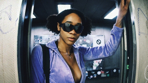 Introspekt has her dark hair in space buns, looking straight at the camera from the doorway of an elevator. She rests her hands on either side, and wears bejewelled sunglasses, a purple velour zip up hoodie, a shoulder bag and and blue jeans. 