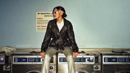 Photo of Paula Tape in a leather jacket, silver trousers and sunglasses. She's sitting on a washing machine in a laundrette