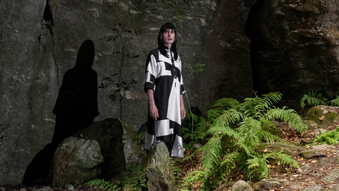 Rrose standing in front of a cliff-like rock structure in a forest, next to a bush of ferns. They are wearing a long black and white dress, a long black wig and make-up. Their shadow is reflected on the wall of the rock 