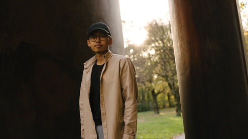 Sam Goku standing in a beige coat and baseball cap in front of a leafy park