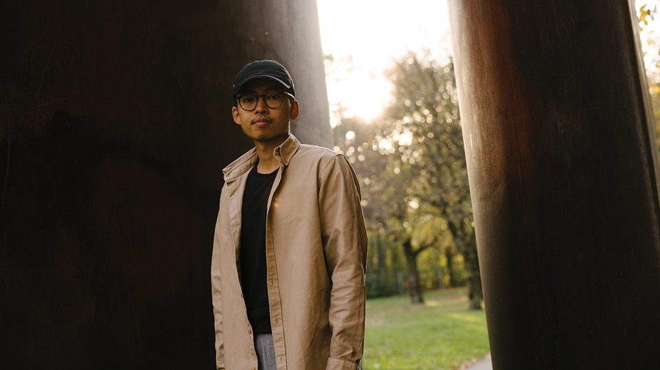 Sam Goku standing in a beige coat and baseball cap in front of a leafy park