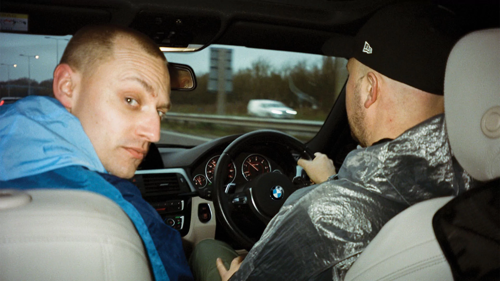 Ed of Overmono looks in toward the camera in the back seat of a car while Tom drives, by Rollo Jackson