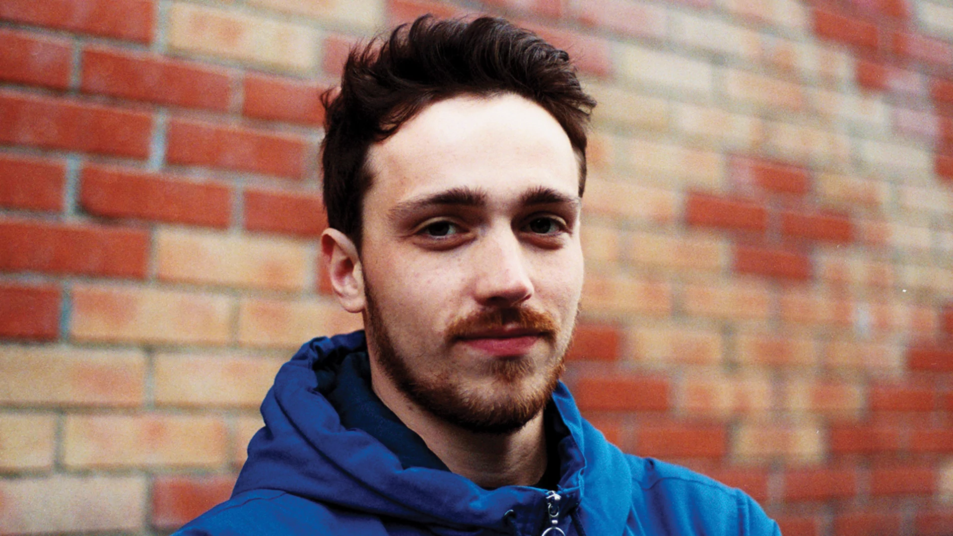 Berzingue in a blue jacket leaning against a patterned red and off-white brick wall