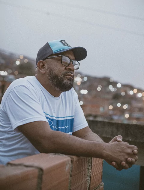 DJ Babatr looking over a wall out at city of Caracas at dusk. He's wearing a white t-shirt, sunglasses and a cap